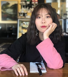 Young Asian woman with dark hair past just her shoulders looks intently at the camera; head resting on her left hand, left elbow resting on a table. Black and pink top. Chairs and pictures behind her.