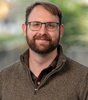 White man with short brown hair, moustache and beard and glasses looks comfortably at the camera. He has a brown sweater on and there is blurred greenery behind him.