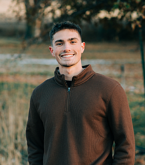 Young white male with short dark hair and a brown top smiles confidently; in the background is water, trees, and grass