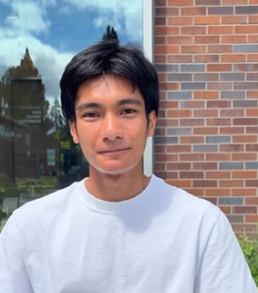 Young Malaysian man with short black hair and a white T-shirt smiles into the camera; a window and brick wall behind him.
