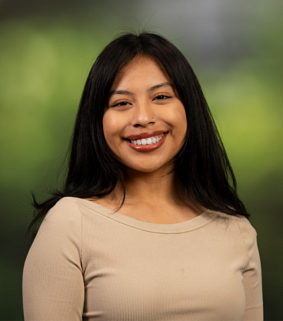 Young woman with long dark hair, wearing a light brown shirt and smiling broadly at the camera