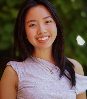 Young Asian woman with long light brown hair and a lilac shirt smiles; greenery behind her