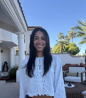 Young woman with long brown hair wearing a white shirt and dark pants; behind her is blue sky, palm trees, and a white building