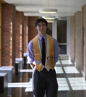 Young man standing in a lilac button-up shirt, dark tie and trousers with two graduation stoles