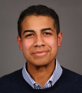 Young man with short brown hair wearing a button down shirt and blue sweater looks steadfastly at the camera