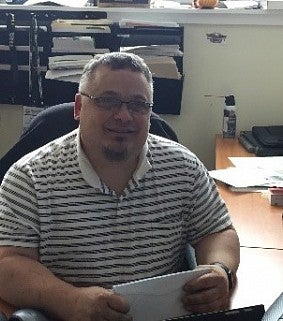 A man with glasses and a goatee sits at a desk; papers are filed on the wall behind