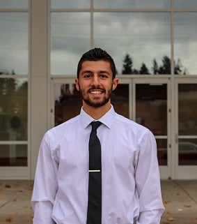 Man with short dark hair, mustache and beard, wearing a white shirt and dark tie smiles at the camera