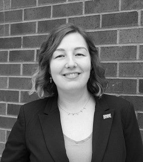 Black and white picture of a woman with short hair, dark jacket and earrings in front of a brick wall