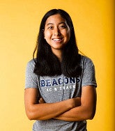Woman with long black hair and folded arms, wearing black pants and a grey t-shirt that reads "Beacons XC & track", smiles in front of a yellow background