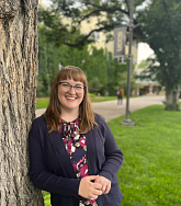 Woman with long brown hair and glasses smiles. She is leaning against a tree; grass, trees, and a walkway visible behind. 