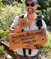 Smiling man wearing a cap and sunglasses holds a sign saying: California/Oregon border