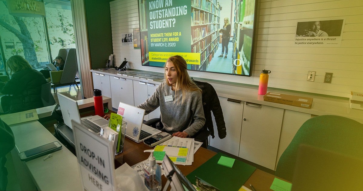 Academic advising receptionist sits at desk helping students check in for drop in advising