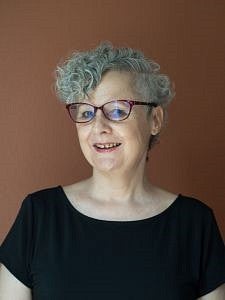 Betsy Wheeler headshot. Betsy is smiling, has short curly grey hair, red glasses, and is wearing a black shirt.