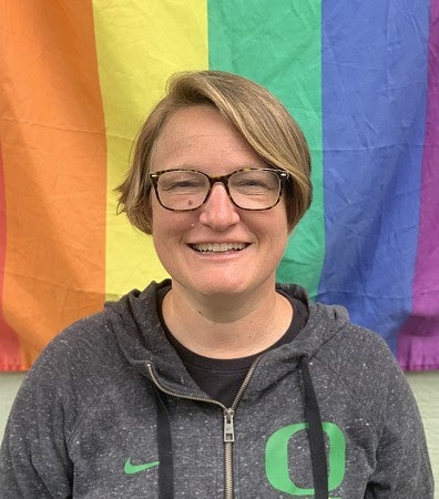 Katie Jacobson smiling with short blonde hair, glasses, wearing a grey UO sweatshirt, and standing in front of a rainbow flag.