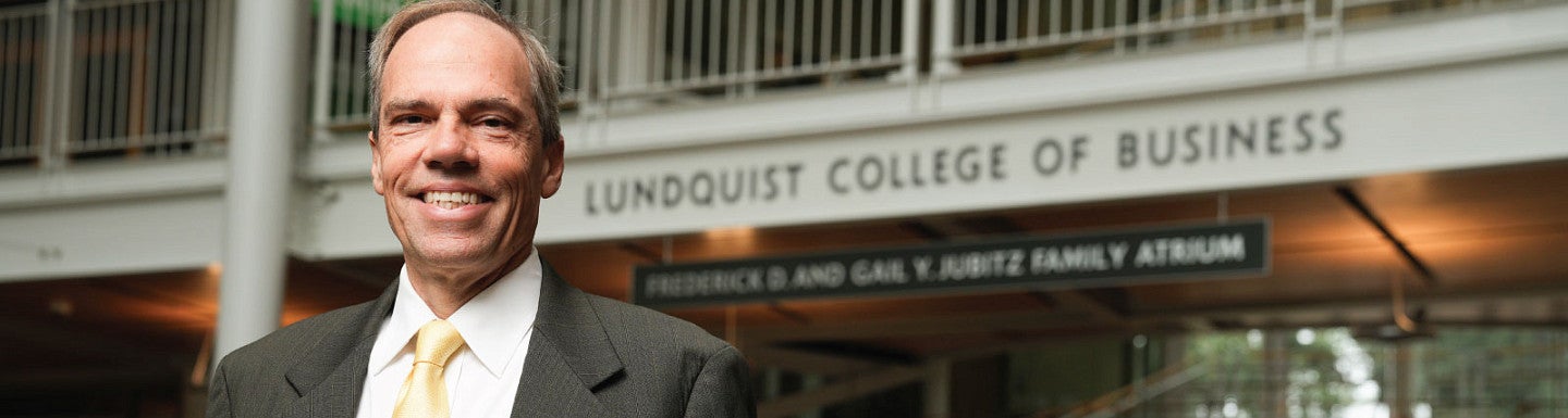 Head shot photo of Bruce Blonigen standing in front the the Lundquist College of Business sign in the Lillis Business Complex atrium