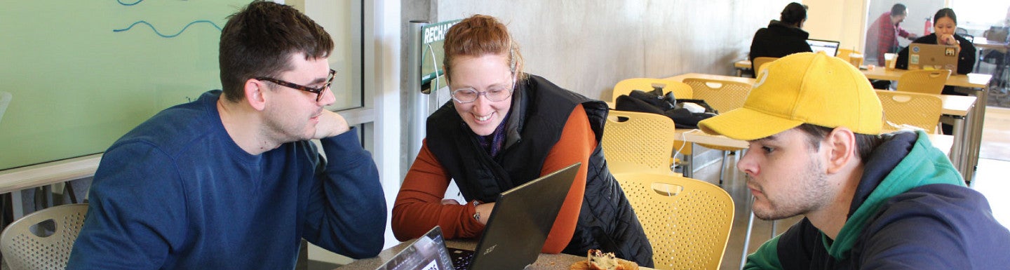 Three students sitting together at a table compare notes on their laptops.