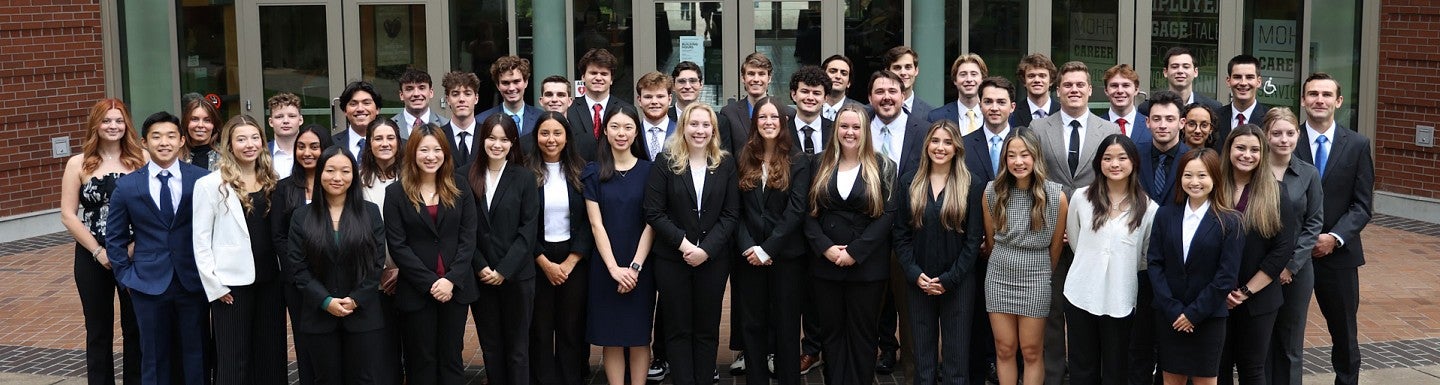A group of honors students stands gathered for a photo in front of the north entrance to the Lillis Business Complex