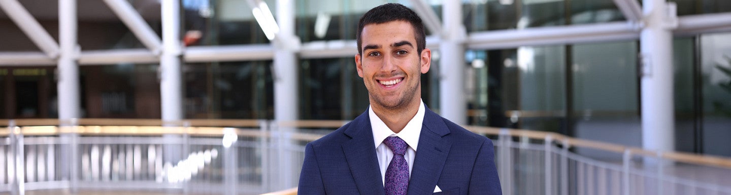Young man in a navy suti with a purple tie stands inside a well-light atrium. windows and a railing can be seen behind him.