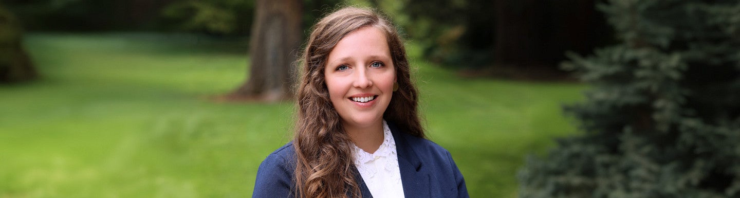 Young women smiles wearing a navy blazer and white shirt. Background is lush and forested.