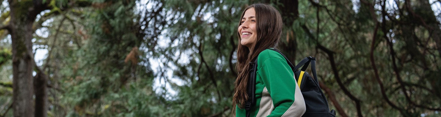 Haylie is looking out into the distance with a backpack over her shoulder. There are pine trees in the background.