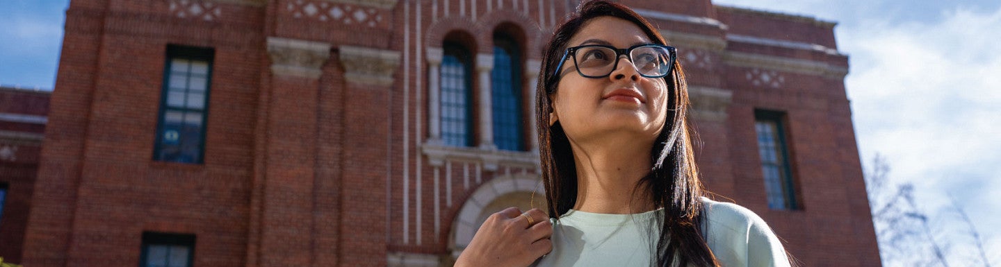 Iffat Rabbani looks up at blue sky above her. A brick building on UO campus is behind her.
