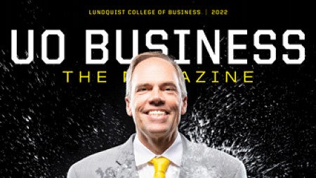 A man wearing a gray suit with a yellow tie is standing against a black backdrop and being splashed with water.