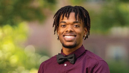 Photo of Jurell Scott smiling at the camera while wearing a maroon shirt with matching bow tie while standing in front of a beautiful bokeh background of colors from campus in spring