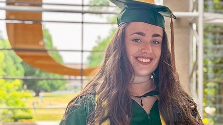 Jorinde van Klinken, wearing commencement regalia, stands in the atrium of the Lillis Business Complex against a backdrop of the giant O on the building's south entrance