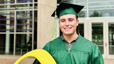 Alex La China, wearing commencement regalia, stands in front of the south entrance to the Lillis Business Complex.