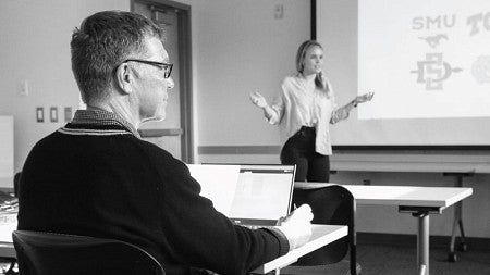 Bob Mighell sits at a classroom table and watches a student make a presentation