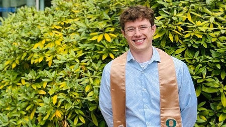 Brett Filuk, MActg ’24, wearing his Master of Accounting graduation sash draped over his shoulders, stands outside the south entrance to the Lillis Business Complex.
