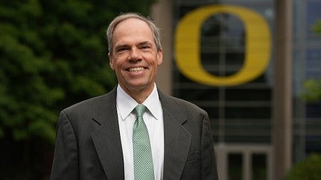 Bruce Blonigen stands in front of the famous O on the south facade of the Lillis Business Complex while wearing a suite and tie and smiling at the camera