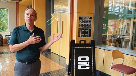 In a screencap from a video, Bruce Blonigen, standing outside the Lundquist Center for Entrepreneurship within the Lillis Business Complex, gestures to the entrance to the center behind him. A white outlined arrow has been overlaid onto the image and is pointing to the center's name above its doors.