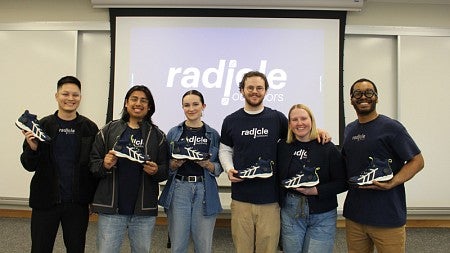 SPM student team Radicle poses for a group photo at the front of the classroom; each member holds their product up in front of themselves