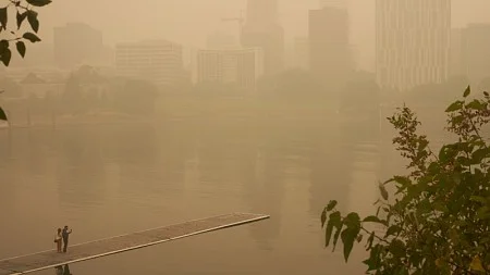 Two people standing on a floating dock look out over a smoggy, smoke-shrouded downtown Portland