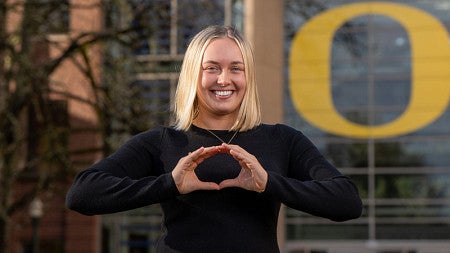 Student Stella Rabb makes the -O- symbol with her hands as she stands outside the south entrance of the Lillis Business Complex on a sunny day