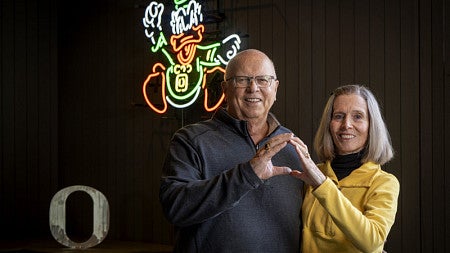 Don (left) and Nancy (right) Smith stand in front of a neon design of the Oregon Duck mascot and make an -O- symbol together with their hands