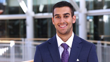 Young man in a navy suti with a purple tie stands inside a well-light atrium. windows and a railing can be seen behind him.