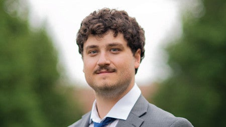 Young man with brown curly hair and a mustach smiles wearing a light gray suit, white shirt, and blue tie. portrait is overlaid on a green and yellow background. 