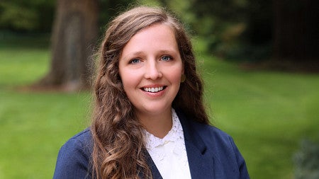 Young women smiles wearing a navy blazer and white shirt. Background is lush and forested.