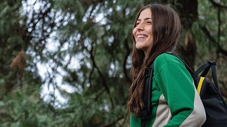 Haylie is looking out into the distance with a backpack over her shoulder. There are pine trees in the background.