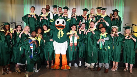 SPM students, all wearing commencement regalia, make the -O- symbol with their hands as they pose for a group photo with the UO Duck mascot