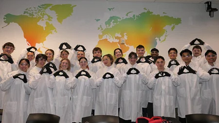 A group of students, dressed in lab coats and gloves, make the -O- symbolk with their hands