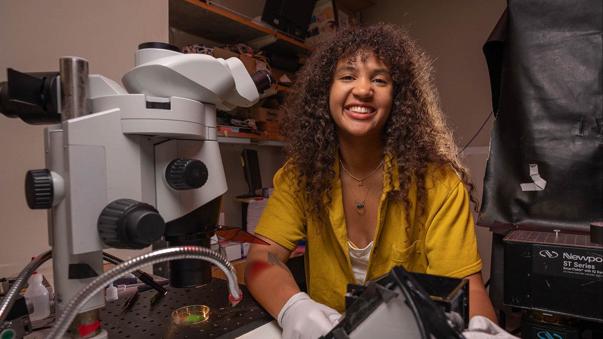 a person beside a microscope who is smiling with long curly hair 