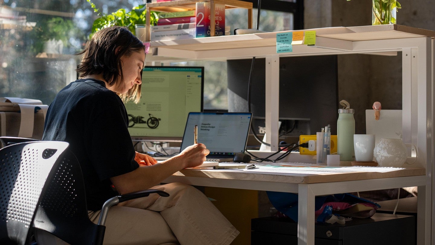 A woman works at a desk with a monitor and a laptop, sun streaming in through the window.