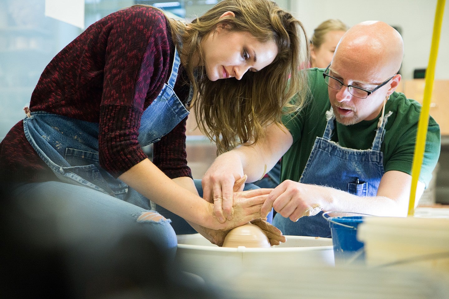 A female student receive instruction on using a pottery wheel from a male instructor.