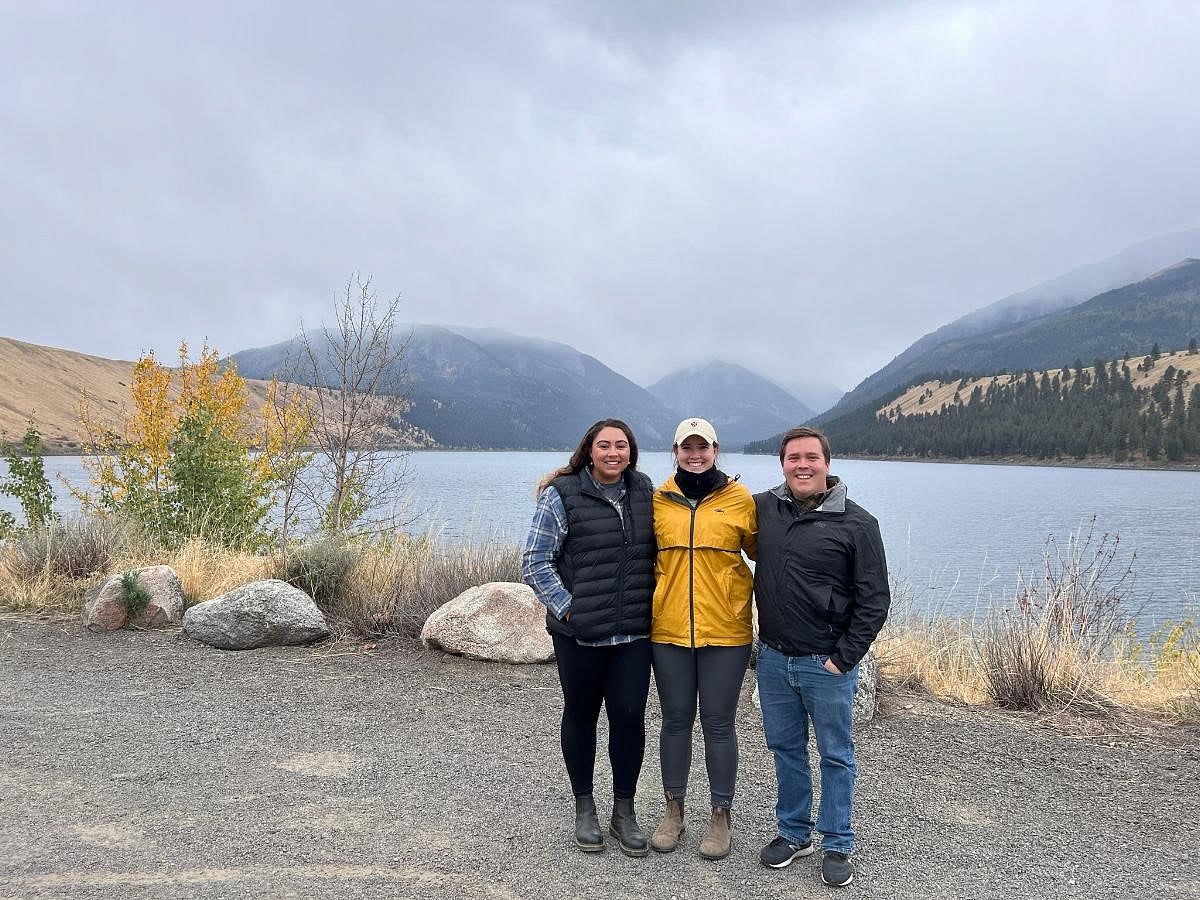 Photograph of three individuals in front of a landscape of hills and water.