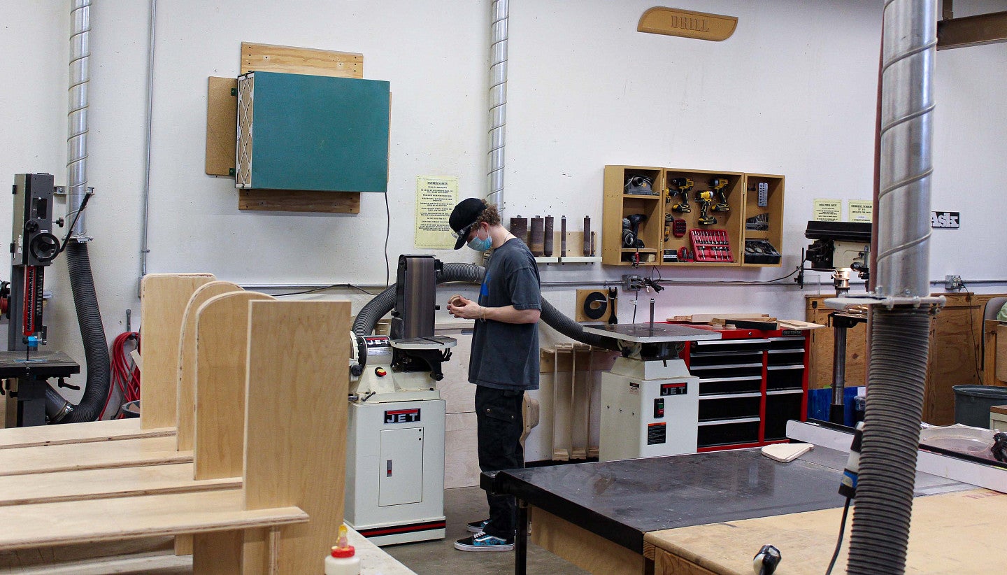 Trent in the woodshop looking over a small wooden piece he has made. What the piece is, is unclear.