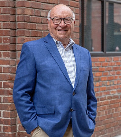 Photograph of Robert Arrigoni. Shows an older man with glasses in a nice, business casual, blue suit jacket, khakis, and a blue and white patterned button up shirt.