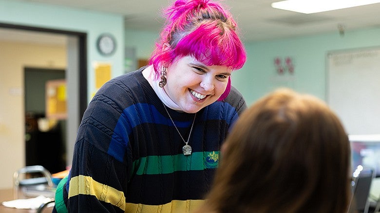 An individual with pink hair speaking to another individual who is sitting